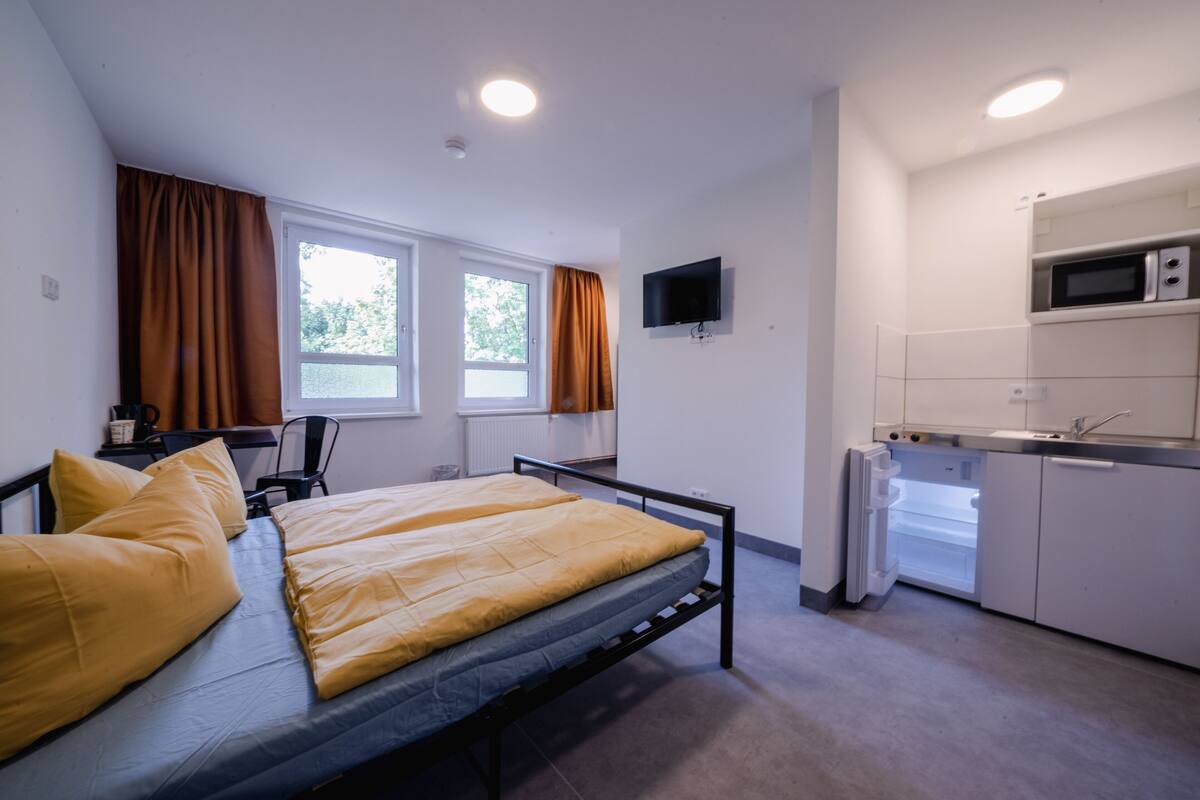 Ein gemütlicher Raum mit Bett, Schreibtisch, Stuhl, Fernseher und einer Küchenzeile.