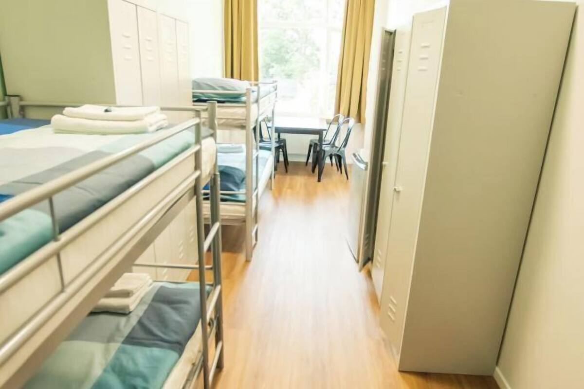 Ein Schlafsaalzimmer mit Etagenbetten, einem Schreibtisch, Stühlen und einem Fenster mit Vorhängen.