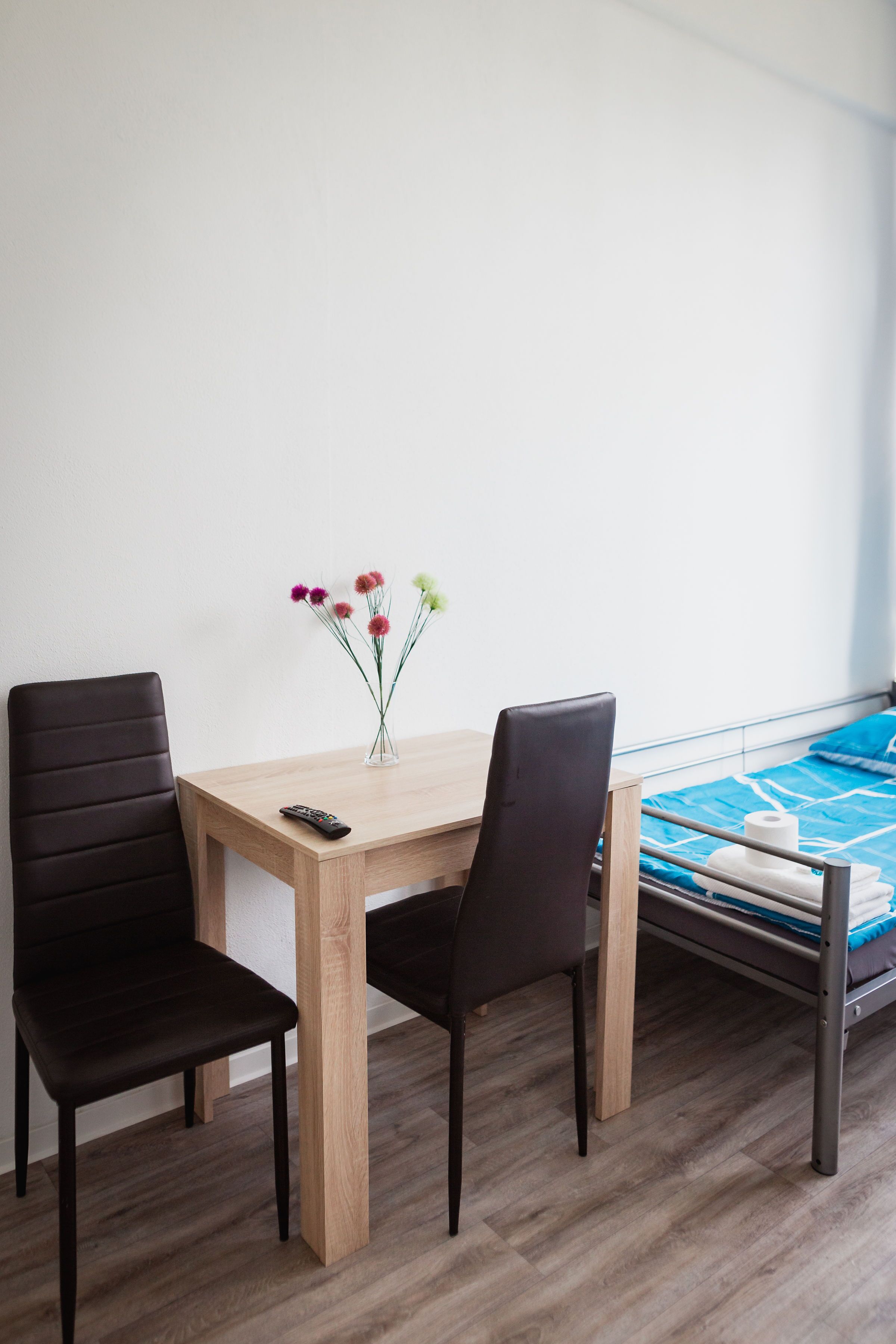 Una habitación con una mesa de madera, dos sillas, un jarrón pequeño con flores, una cama con una manta azul y un control remoto sobre la mesa.