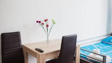 Una habitación con una mesa de madera, dos sillas, un jarrón pequeño con flores, una cama con una manta azul y un control remoto sobre la mesa.