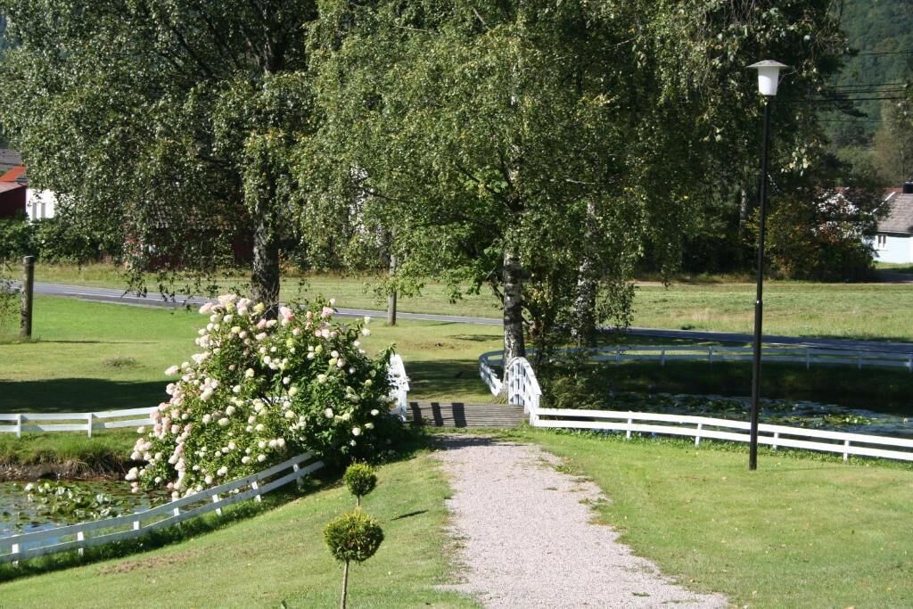 A pathway leading to a white bridge over a pond, surrounded by lush greenery and a white fence.