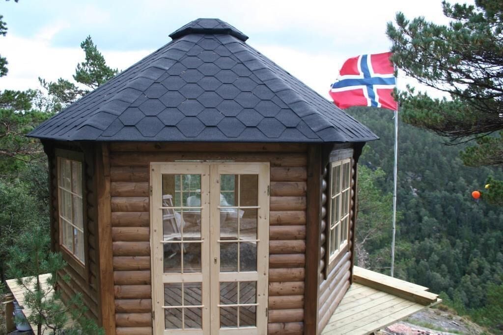 A wooden cabin with a hexagonal roof, a flagpole with a flag, and a view of a forested mountain area.