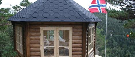 A wooden cabin with a hexagonal roof, a flagpole with a flag, and a view of a forested mountain area.