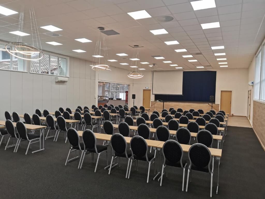 Ein Konferenzraum mit Tischen und Stühlen, einer Bühne mit einem Bildschirm und Pendelleuchten.