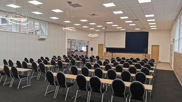 Ein Konferenzraum mit Tischen und Stühlen, einer Bühne mit einem Bildschirm und Pendelleuchten.