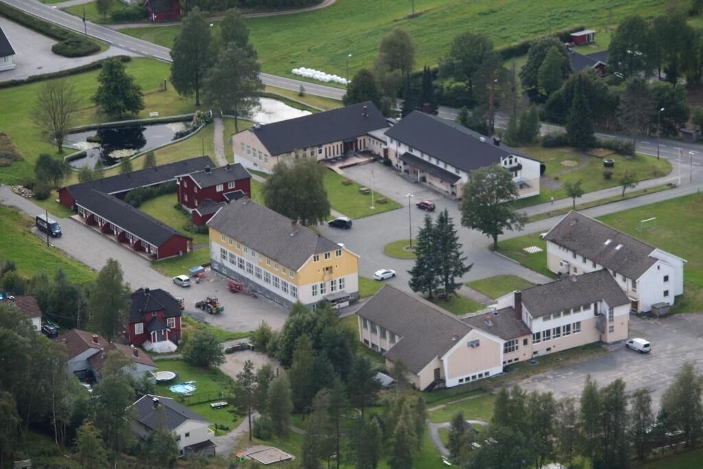 Ett flygbildsbild av ett bostadsområde med flera hus och en stor gul byggnad, troligen en skola eller ett samhällscentrum, omgivet av gröna fält och träd.