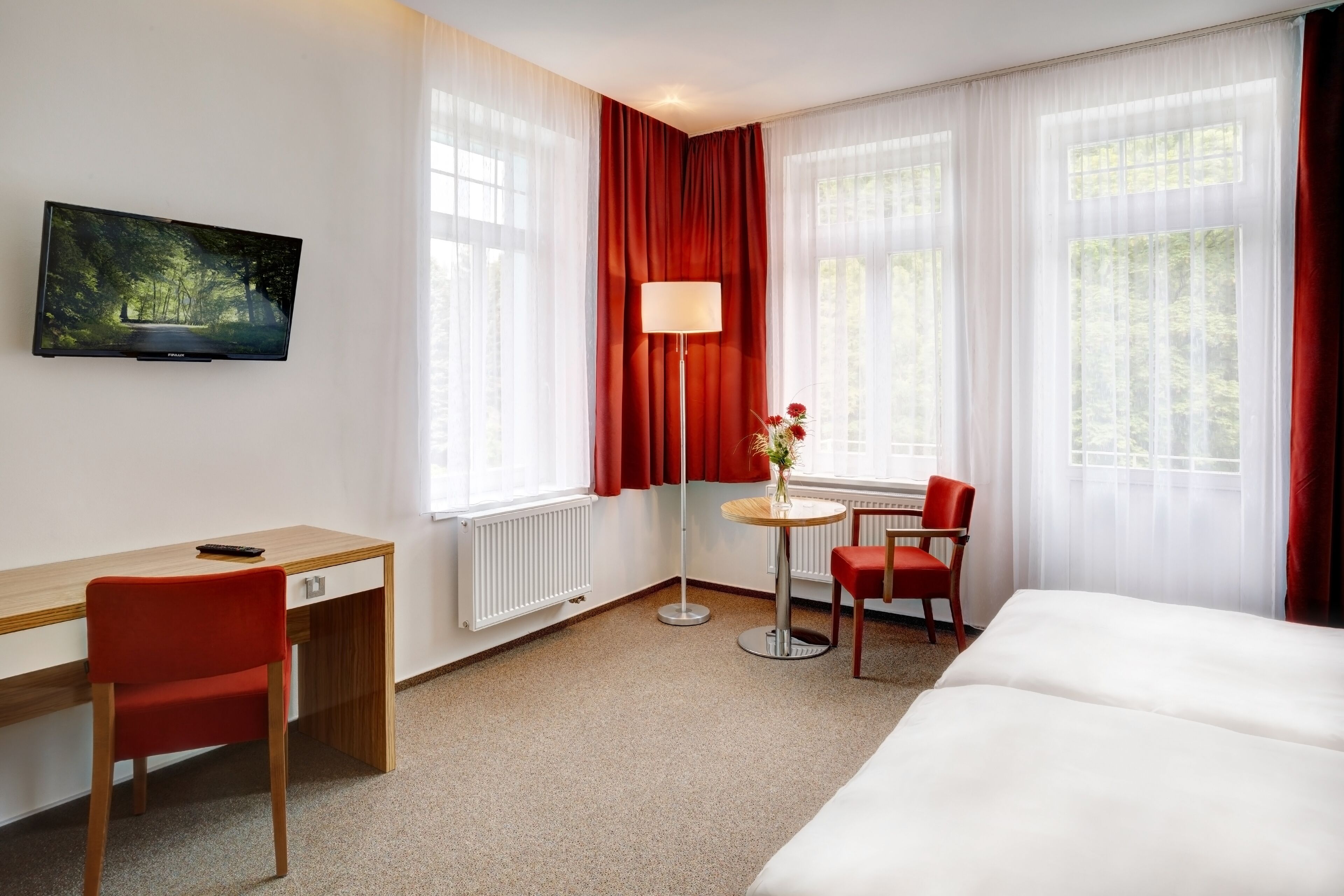 Ein Hotelzimmer mit Bett, Schreibtisch, Stuhl, einem kleinen Tisch mit Lampe und einem Fernseher an der Wand.