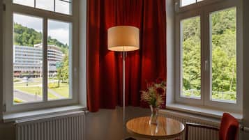 A hotel room with a large window, a red curtain, a floor lamp, a small table with a vase of flowers, and a chair.