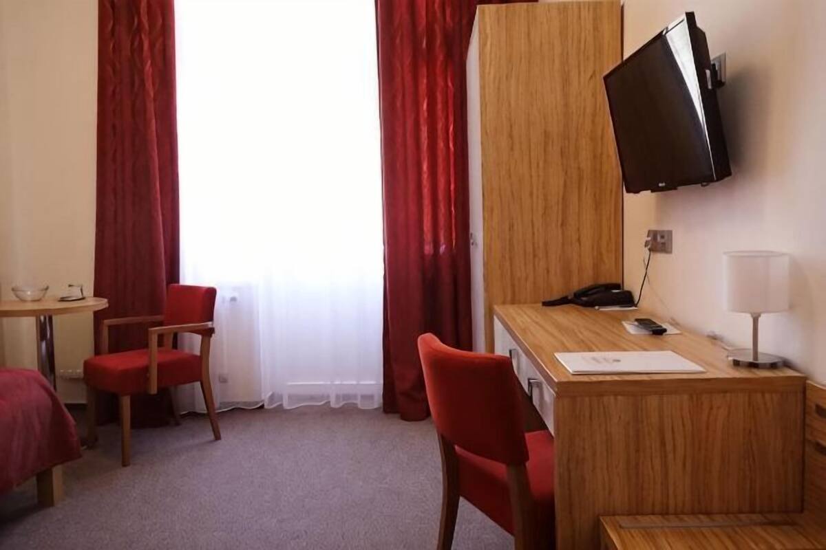 A hotel room with a desk, two red chairs, a bed, a TV, and a window with curtains.