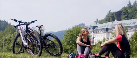 Dos personas sentadas sobre la hierba cerca de una bicicleta de montaña, con un edificio grande al fondo.