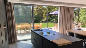 Une cuisine moderne dotée d’un grand îlot central et de portes-fenêtres coulissantes donnant sur une terrasse extérieure aménagée avec du mobilier en bois.