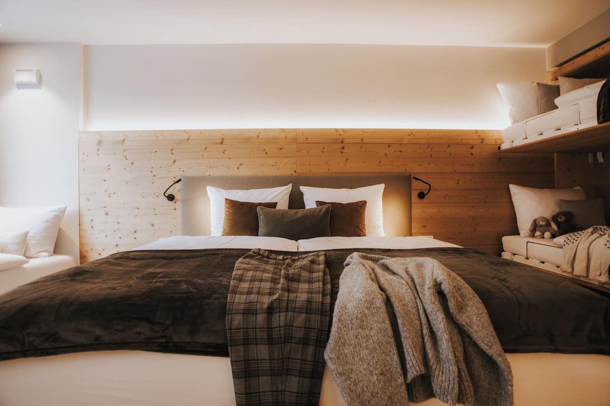 Une chambre à coucher avec une tête de lit en bois, un lit recouvert d’une couverture foncée, une table de chevet avec une lampe et une petite étagère ornée d’objets décoratifs.