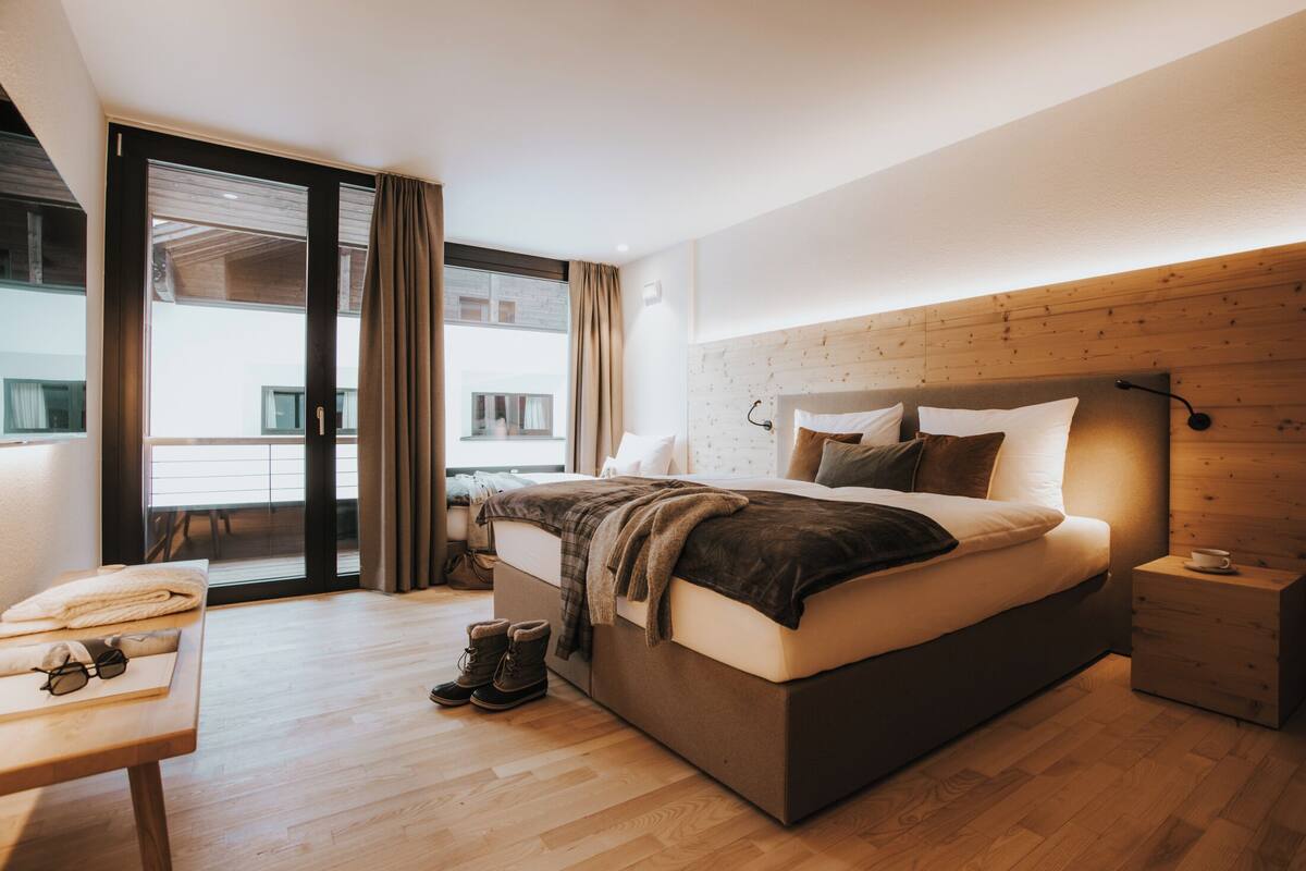 Une chambre moderne avec un grand lit, une tête de lit en bois, une table de chevet et une vue sur l’extérieur grâce à une porte vitrée.