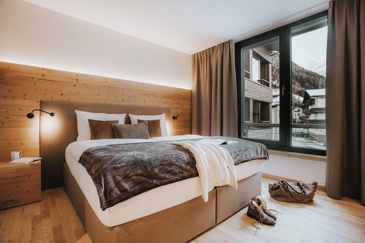 Une chambre moderne avec une tête de lit en bois, un grand lit recouvert d’une couette marron et blanche, une table de chevet avec une tasse, et une vue sur une montagne enneigée par la fenêtre.