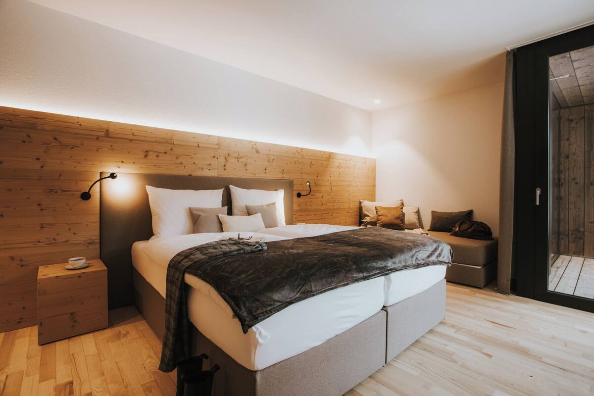 Une chambre moderne avec une tête de lit en bois, un grand lit recouvert de draps blancs, une table de chevet avec une tasse, et une vue à travers une porte vitrée.