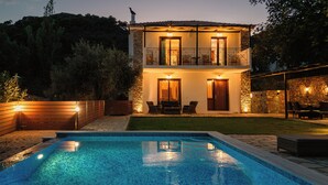 Una casa de dos pisos con piscina, mobiliario exterior y un muro de piedra.