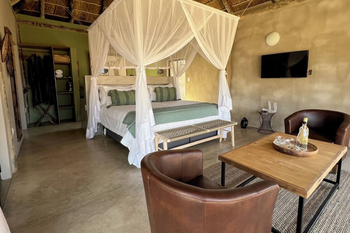A room with a bed under a canopy, a small table with a bottle, a chair, and a TV on the wall.