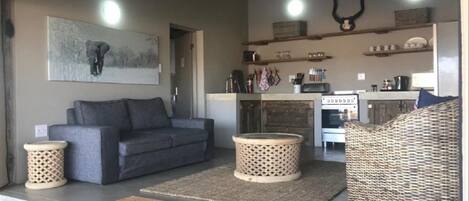 A cozy living area with a grey sofa, a wicker chair, and a kitchen in the background.
