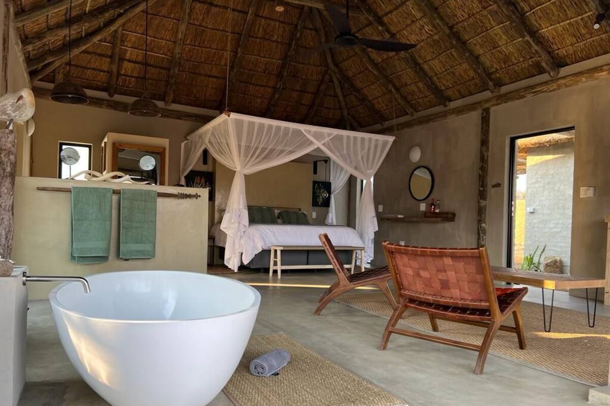 A room with a large white bathtub, a wooden chair, a bed with a canopy, and a thatched ceiling.