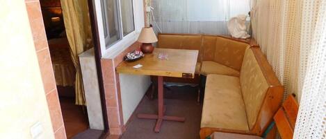 Una habitación de esquina con una mesa de madera, un banco acolchado, una lámpara pequeña y una ventana con cortinas.