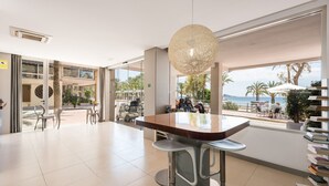 Um lobby de hotel moderno com uma janela ampla que oferece vista para a praia e palmeiras. Há banquetas de bar e uma mesa com cadeiras.