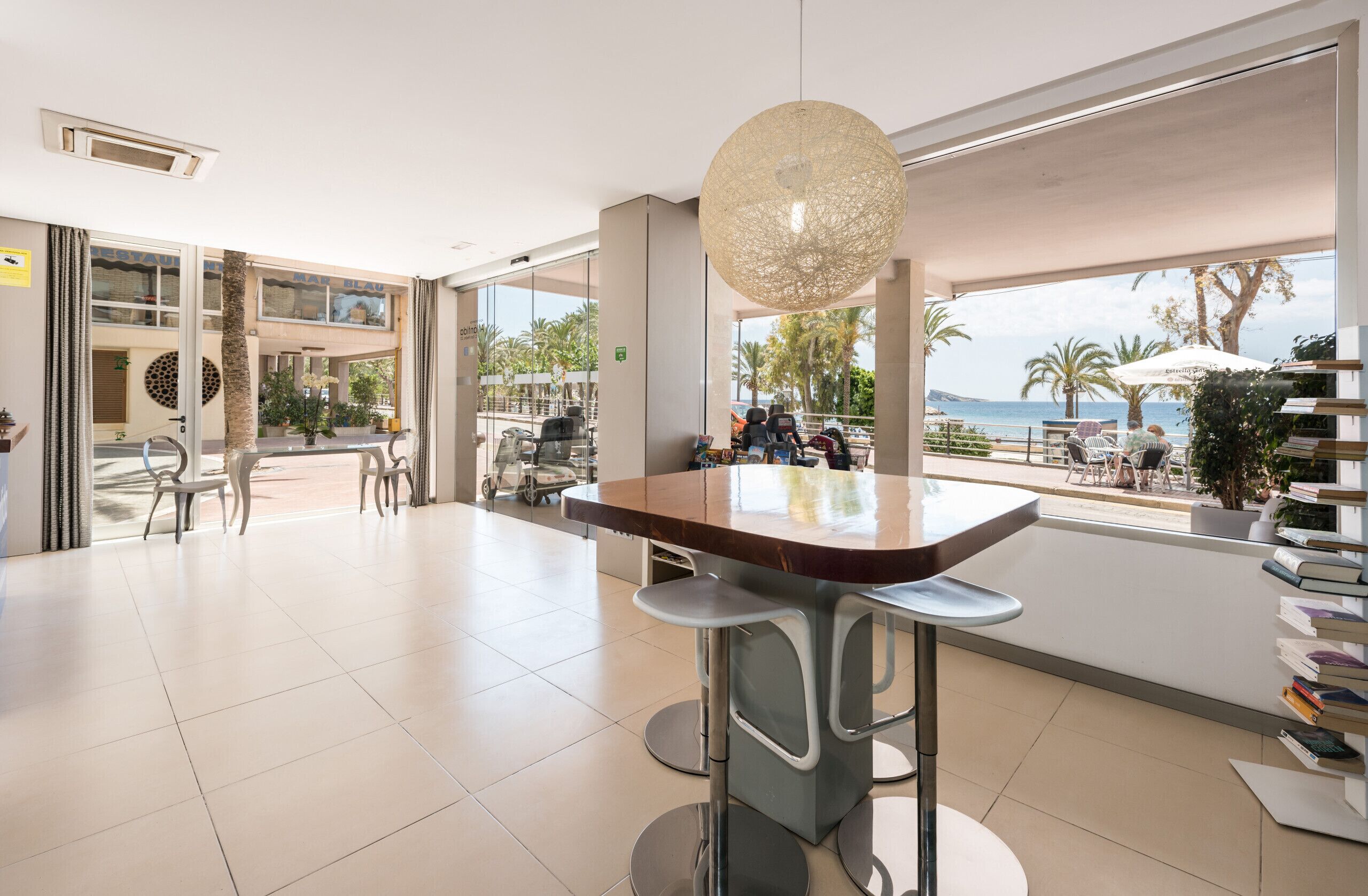 Recepció d'hotel moderna amb una finestra gran que ofereix vistes a la platja i palmeres. Hi ha tamborets i una taula amb cadires.