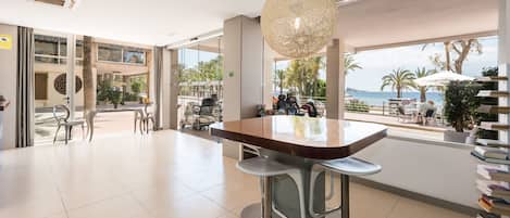 Un hall d’hôtel moderne doté d’une grande fenêtre offrant une vue sur la plage et les palmiers. On y trouve des tabourets de bar et une table avec des chaises.