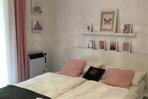 A bedroom with a bed, pillows, a bedside table, and a heater. There are wall decorations and a window with curtains.
