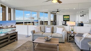 A spacious living room with a large window, a ceiling fan, and a view of the ocean. - Sandpiper Condominiums 304, Panoramic Gulf Views w/ direct access to Mustang Island/Port A beaches. (Port Aransas)