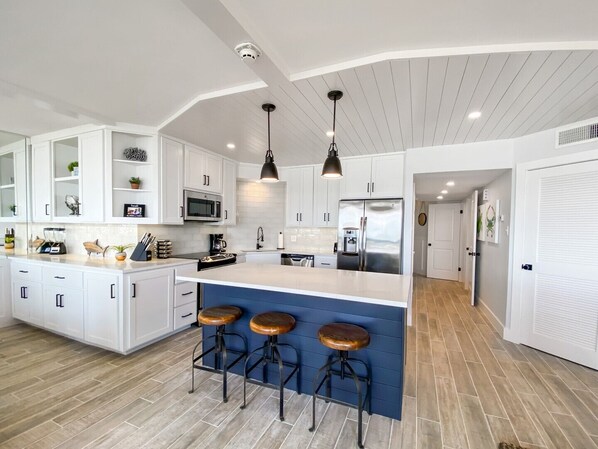 A modern kitchen with white cabinetry, a central island with stools, stainless steel appliances, and pendant lighting. - Sandpiper Condominiums 304, Panoramic Gulf Views w/ direct access to Mustang Island/Port A beaches. (Port Aransas)