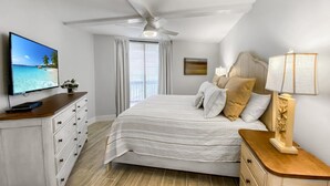A bedroom with a bed, a TV, a ceiling fan, a nightstand, and a window with curtains. - Sandpiper Condominiums 304, Panoramic Gulf Views w/ direct access to Mustang Island/Port A beaches. (Port Aransas)