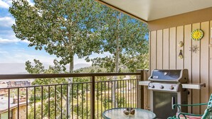 A balcony with a grill, a table, and chairs, overlooking a cityscape and a tree. - Summer in Snowmass! Spacious Condo w/ Sleeping Den, Pool, Hot Tub, Balcony W/ Grill, FP & Parking (Snowmass Village)