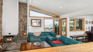A spacious living room with a stone fireplace, green leather sofa, and large windows offering a view of the surrounding area. - Summer in Snowmass! Spacious Condo w/ Sleeping Den, Pool, Hot Tub, Balcony W/ Grill, FP & Parking (Snowmass Village)