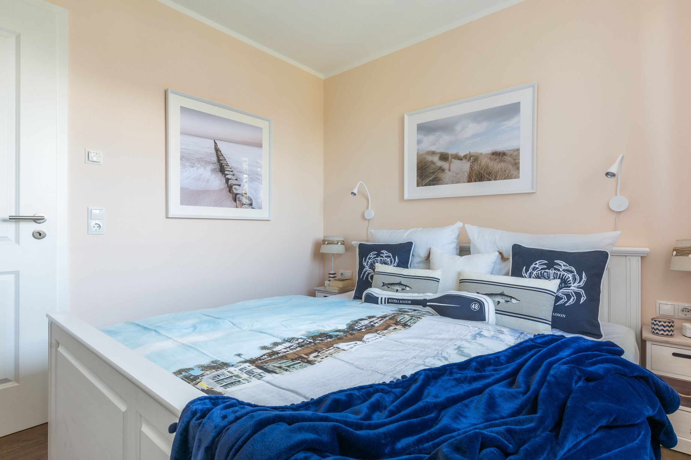 A bedroom with a bed, two bedside tables, a nightstand, a wall-mounted lamp, and two framed pictures.
