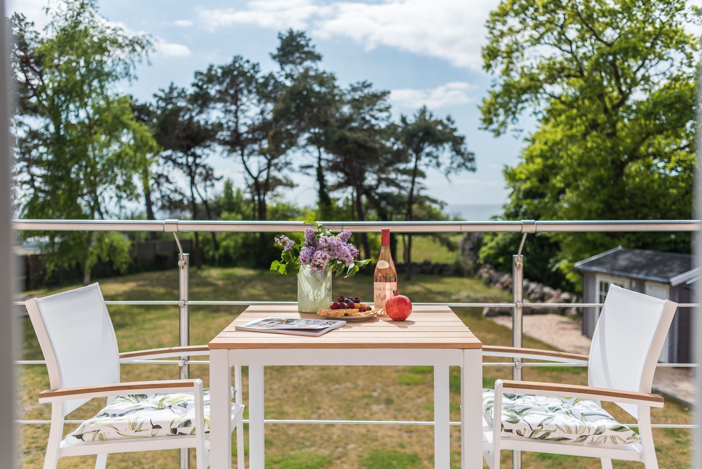 Un balcon avec une table dressée pour deux personnes, agrémenté d’un vase de fleurs, d’une bouteille et d’une assiette de nourriture.