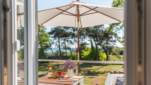 Un balcone con tavolo in legno, sedie bianche, un ombrellone bianco e vista su alberi e uno specchio d'acqua.