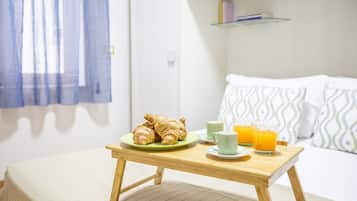 A bed with a white and patterned pillow, a bedside table with croissants, a green plate, two mugs, and two glasses of orange juice.