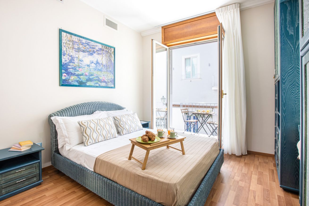 A bedroom with a bed, a bedside table with books, a painting on the wall, and a balcony with a table and chairs.