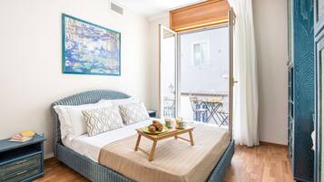 A bedroom with a bed, a bedside table with books, a painting on the wall, and a balcony with a table and chairs.