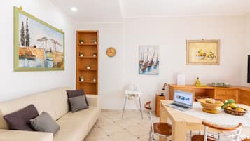 A living room with a sofa, a dining table with chairs, a laptop, and a painting on the wall.