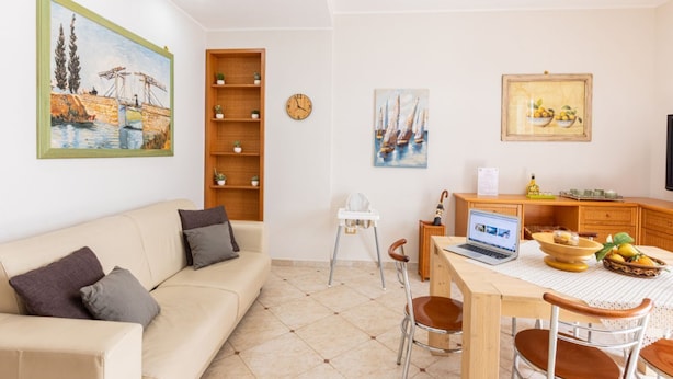 A living room with a sofa, a dining table with chairs, a laptop, and a painting on the wall.