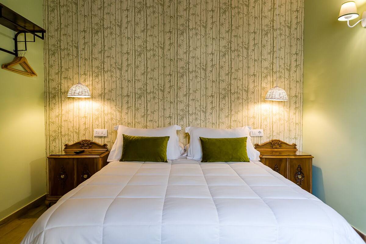 Una cama bien hecha con ropa blanca y almohadas verdes, flanqueada por dos mesitas de noche con lámparas, frente a una pared con papel tapiz de bambú vertical.