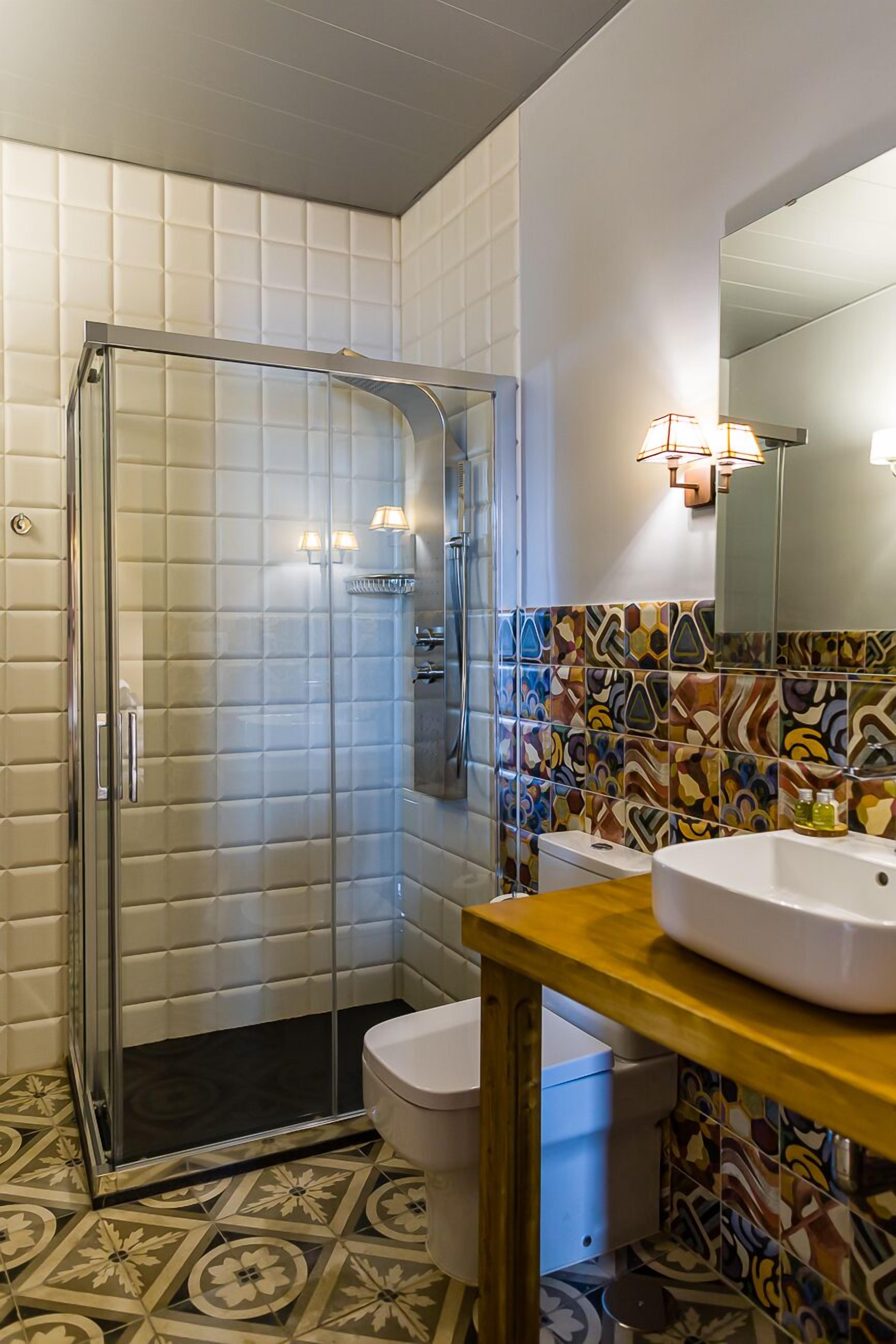 Een badkamer met een douchecabine van glas, een toilet en een wastafel met spiegel erboven. De wanden zijn betegeld met verschillende patronen, en er is een houten aanrecht onder de wastafel.