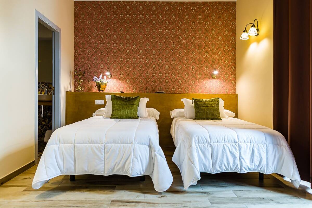 Habitación de hotel con dos camas, papel tapiz estampado y una mesita de noche con un florero.