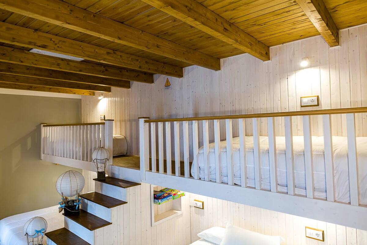 Una escalera de madera lleva a una habitación con literas, barandilla blanca y una estantería con libros.