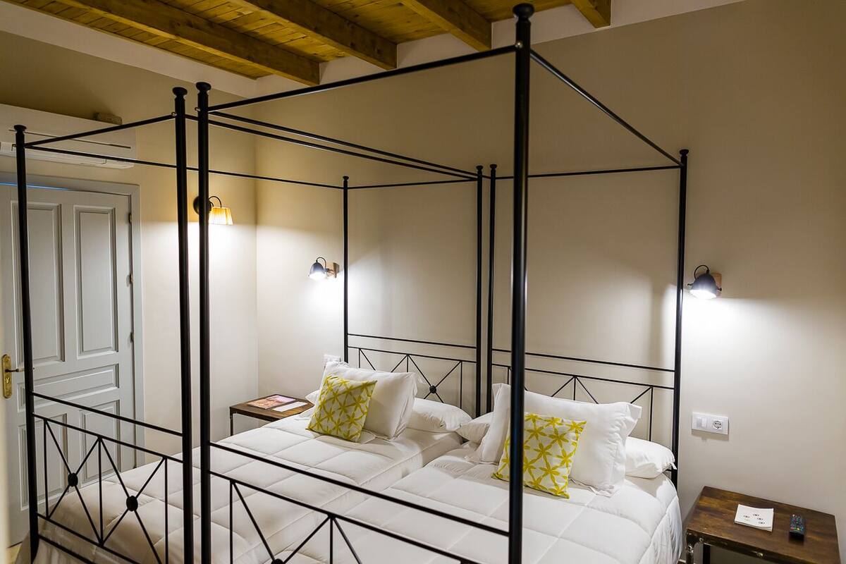 Una cama con dosel con ropa de cama blanca y almohadas estampadas en amarillo, una mesita de noche con lámpara y una pequeña mesita auxiliar de madera.