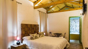Une chambre avec un plafond en bois, un lit avec une literie blanche, une table de chevet avec une lampe et une porte verte donnant sur l’extérieur.