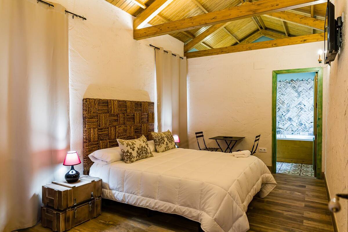 Un dormitorio con techo de madera, una cama con ropa de cama blanca, una mesita de noche con lámpara y una puerta verde que da al exterior.