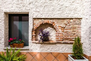 A rustic brick wall with a window, a small shelf with potted plants, and a tiled floor.