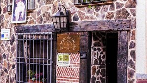 Un bâtiment en pierre doté d’une entrée rustique, d’une enseigne en bois et d’une fenêtre grillagée ornée de plantes en pot.